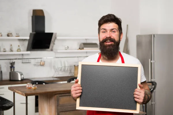 Man bearded hipster red apron stand in kitchen. Kitchen furniture store. Kitchen hacks concept. Clever ways to organize kitchen. Cook hold blank chalkboard copy space. Secret tips. Useful information