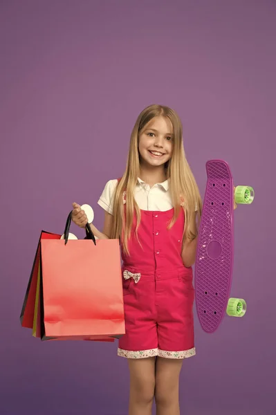 Chica feliz mantenga penny board y bolsas de compras sobre fondo violeta. Sonrisa infantil con paquetes y monopatín. Patinadora sonriendo con bolsas de papel. Compras y venta. Deporte y hobby activo —  Fotos de Stock