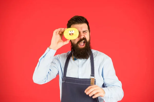 Oublie le régime. Manger le repas. Hipster barbu boulanger tenir beignet glacé sur fond rouge. Café et concept de boulangerie. Doux beignet du boulanger. Homme barbu boulanger dans tablier de cuisine tenir dessert mignon — Photo