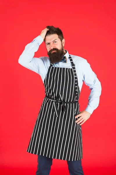 Concept de café moderne. Cuisiner des repas modernes. Homme avec barbe cuisinier tablier hipster. Hipster chef cuisinier fond rouge. Un homme barbu cuisinier. Hipster cuisine maison ou restaurant. Confiant dans sa recette — Photo