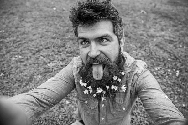 Un type avec des fleurs de chélandine en barbe qui prend des photos de selfie. Hipster avec grimace joyeuse avec la langue sur le visage prenant selfie. L'homme aime le printemps, fond vert, déconcentré. Concept d'humeur joyeuse — Photo