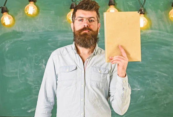 Profesor en anteojos tiene libro en la mano, espacio de copia. El hombre con barba y bigote en la cara tranquila se para en el aula. Concepto de literatura científica. Científico sostiene libro, pizarra en el fondo — Foto de Stock