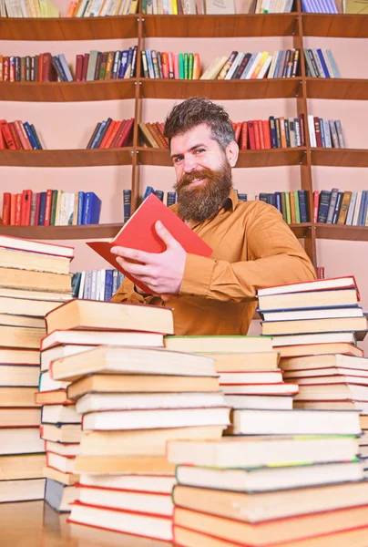 Conceito de educação e ciência. Professor ou estudante com barba estudando na biblioteca entre pilhas de livros. Homem na cara sorridente leitura livro, estantes no fundo — Fotografia de Stock