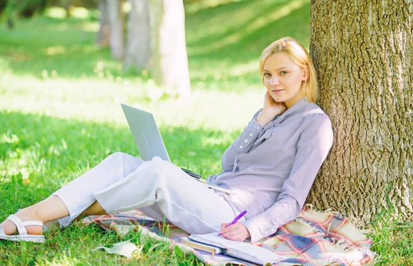Преимущества работы на открытом воздухе. Женщина с ноутбуком работает на открытом воздухе опираясь на ствол дерева. Девушка, работающая с ноутбуком в парке, сидит на траве. Образовательные технологии и интернет-концепция. Природный офис — стоковое фото