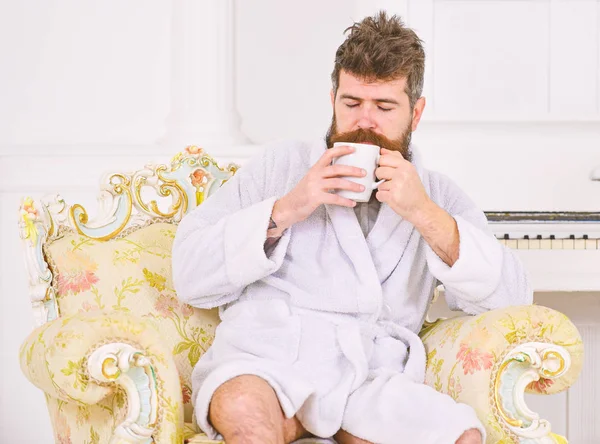 男はバスローブを着て、飲み物コーヒーで眠そうな朝、白い背景に高級ホテルで香りを楽しんでします。ひげを持つ男、口ひげは高級アームチェアに座って朝を楽しんでいます。エリート レジャー概念 — ストック写真