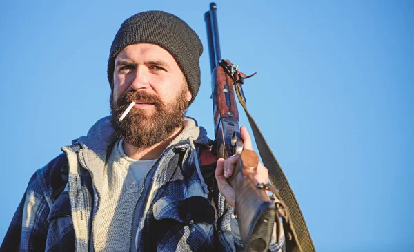 Man brutal bearded guy gamekeeper blue sky background. Brutality and masculinity. Hunter with rifle gun close up. Guy bearded hunter spend leisure hunting and smoking. Hunting masculine hobby concept