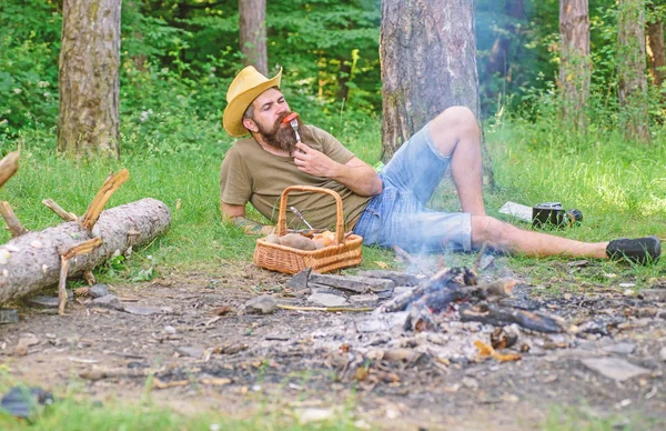 Man bearded hipster lay near campfire after day hiking or gathering mushrooms. Man have snack or picnic in forest. Guy relaxing near campfire and eating snacks. Brutal bearded tourist relaxing