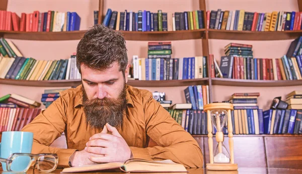 Uomo su libro di lettura di faccia concentrato, studiando, librerie su sfondo. Insegnante o studente con barba che studia in biblioteca. L'uomo siede a tavola con tazza e clessidra. Concetto di autoeducazione — Foto Stock