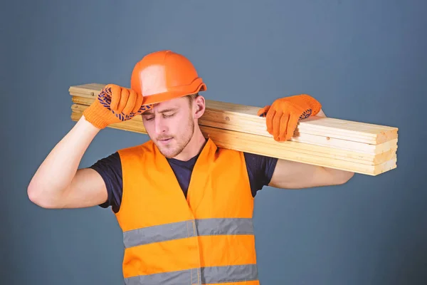 Έννοια ασφάλειας και προστασίας. Ξυλουργός, ξυλουργός, ισχυρό οικοδόμος στο πολυάσχολο πρόσωπο ασκεί ξύλινα δοκάρια στον ώμο. Ο άνθρωπος σε προστατευτικά γάντια κατέχει προστατευτικό σκληρό καπέλο, γκρι φόντο της προσωπίδας — Φωτογραφία Αρχείου
