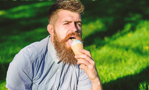 Bearded man with ice cream cone. Delicacy concept. Man with long beard eats ice cream, while sits on grass. Man with beard and mustache on sexy face licks ice cream, grass on background, defocused