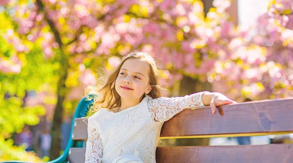 Koncepcja wiosna. Dziewczyna na uśmiechniętą twarz siedzi na ławce, drzewa sakura na rozmytym tłem. Relaks podczas spaceru w parku, w pobliżu wiśniowy kwiat dziewczyna. Słodkie dzieci cieszyć się dzień słoneczny wiosna — Zdjęcie stockowe