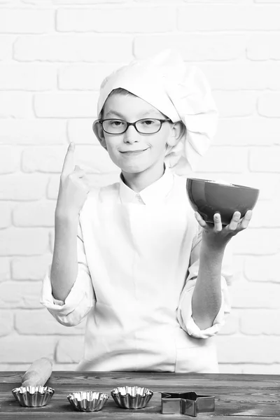 Jeune garçon petit chef cuisinier mignon en uniforme blanc et chapeau sur le visage drôle avec des lunettes debout près de la table avec des moules à rouleau pour gâteaux et tenant bol rouge sur fond de mur de brique — Photo