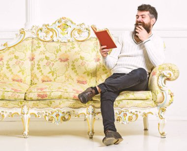 Sakal ve bıyık oturur barok tarzı kanepe, tutar kitap, beyaz duvar arka plan üzerinde olan adam. Adam zevk ile eski kitap okuma. Mizahi edebiyat kavramı. Maço yüz kitap okuma gülüyor üzerinde