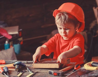 Kask oluşturucu veya repairer, onarımı veya handcrafting olarak oynayan sevimli çocuk. Meşgul yüz oyunlarında evde atölye üzerinde yürümeye başlayan çocuk. Kavram handcrafting. Çocuk çocuk dövülmüş tırnak içine ahşap tahta