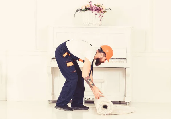 Relocating concept. Courier delivers furniture in case of move out, relocation. Man with beard, worker in overalls and helmet rolling carpet, white background. Loader wrappes carpet into roll