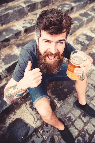 Hombre con barba y bigote sostiene el vaso con cerveza y muestra el pulgar hacia arriba, escaleras de piedra de fondo. Hipster en la cara alegre bebiendo cerveza al aire libre. Un tipo descansando con cerveza fría. Viernes relajar concepto — Foto de Stock