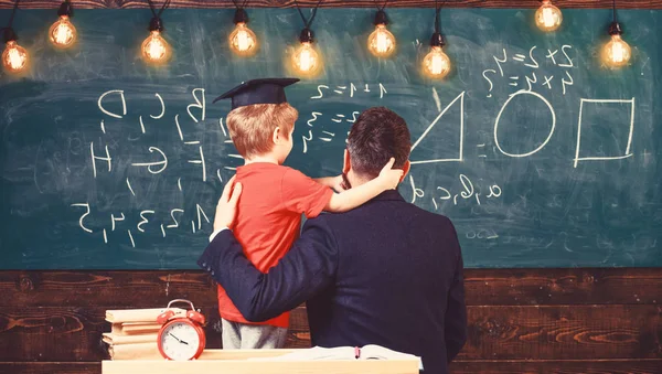 Teacher with beard, father hugs little son in classroom while discussing, chalkboard on background. Best friends concept. Child in graduate cap listening teacher, chalkboard on background, rear view