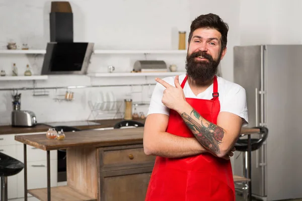 Tienda de muebles de cocina. Cocinar en cocina nueva. Necesito inspiración culinaria. El fin de semana comienza con un delicioso desayuno. Cómo convertir la cocina en casa en hábito. Caballero barbudo hipster delantal rojo pie en la cocina —  Fotos de Stock