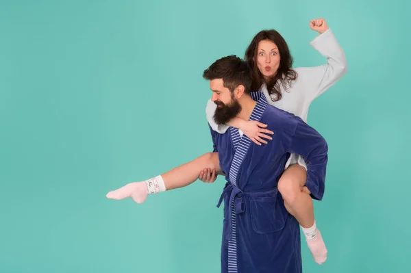 Handsome young man giving his girlfriend piggyback ride. Couple in bathrobes having fun turquoise background. Lets stay at home and have fun. They always have fun together. Close relationship