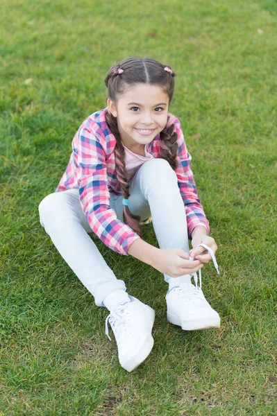 Legare i lacci delle scarpe. Ragazzina bambino trascorrere il tempo libero all'aperto nel parco. Ragazza seduta sull'erba nel parco. Bambino godere di primavera tempo soleggiato mentre seduto al prato nel parco. Idea primaverile. Parco e giardino — Foto Stock