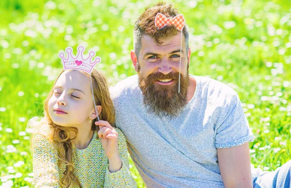 Familia pasar el ocio al aire libre, jugar juegos femeninos. Papá y su hija se sientan en la hierba en Grassplot, fondo verde. Niño y padre posando con atributos de cabina de fotos de corona y arco. Concepto de paternidad — Foto de Stock