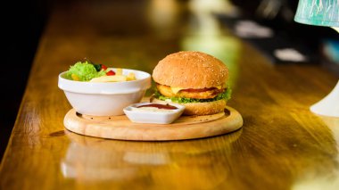 Delicious burger with sesame seeds. Burger with cheese meat and salad. Fast food concept. Burger menu. High calorie snack. Hamburger and french fries and tomato sauce on wooden board. Cheat meal clipart