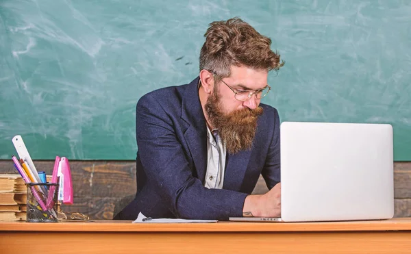 Escribiendo informe escolar. Profesor de la escuela escribiendo informe portátil. Profesor sentarse en el escritorio con el ordenador portátil. De vuelta al concepto escolar. Maestro barbudo hipster con anteojos sentarse en el aula pizarra fondo —  Fotos de Stock