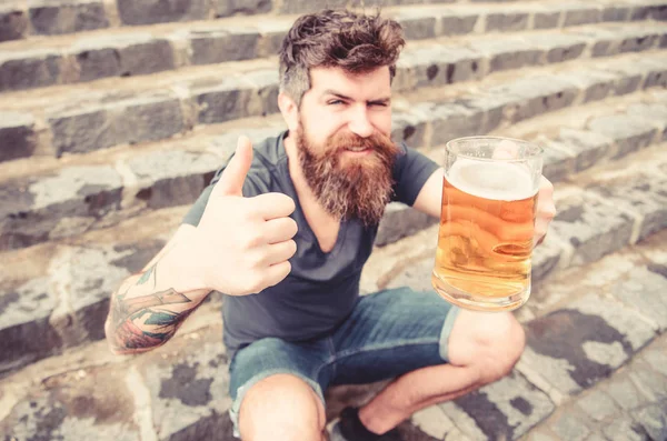 Człowiek z broda i wąsy posiada szkło z piwa i pokazuje kciuk w górę, schody kamienne podłoże. Facet po odpoczynku z piwem, rozmyte. Hipster na wesołą twarz pije piwo odkryty. Piątek zrelaksować się koncepcja — Zdjęcie stockowe