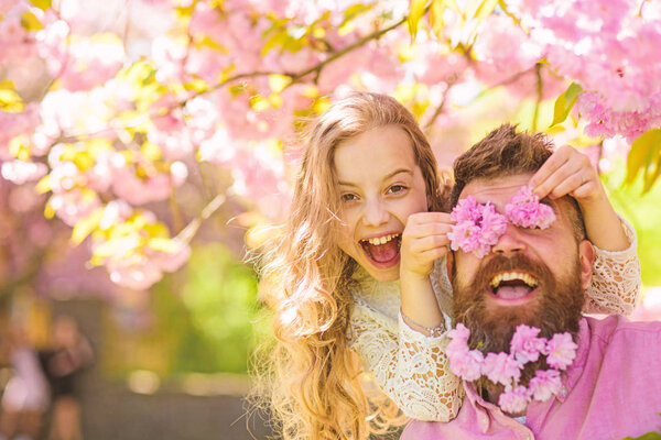 Child and man with tender pink flowers in beard. Girl with dad near sakura flowers on spring day. Father and daughter on happy face play with flowers as glasses, sakura background. Family time concep