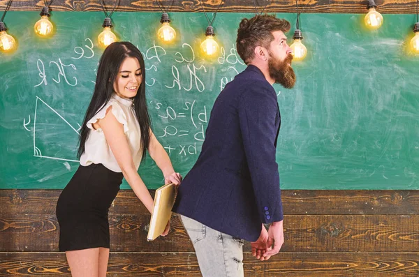 Chica en la cara sonriente divirtiéndose mientras castiga a profesor. Estudiante dama abofeteando a profesores nalgas con libro. Concepto de juegos de rol. Hombre con barba castigado por estudiante sexy, pizarra en el fondo —  Fotos de Stock