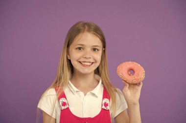 Do you want bite. Girl smiling face holds pink donut in hand, violet background. Kid smiling girl ready to bite donut. Snack concept. Child can not wait to eat sweet donut, copy space clipart