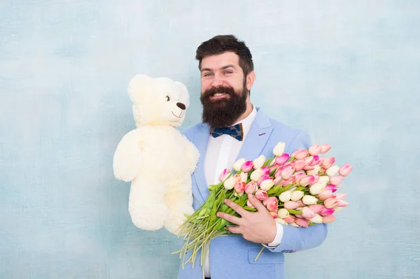 Man bearded gentleman suit bow tie hold teddy bear and bouquet. Happy and in love. Gentleman making romantic surprise for her. Gentleman romantic gift. Birthday greetings. Happy birthday my love