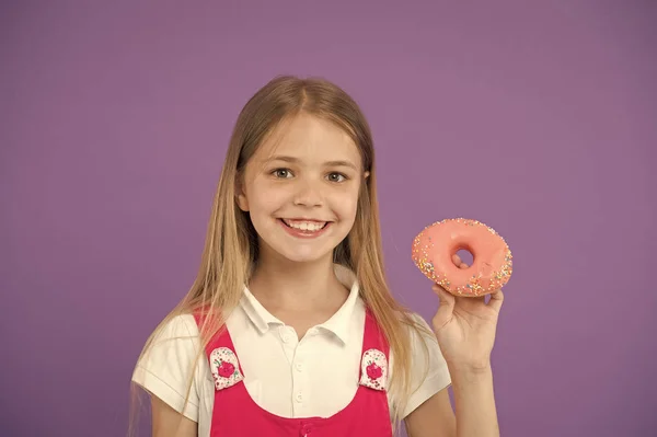 Queres morder? Menina sorridente rosto segura rosa donut na mão, fundo violeta. Garota sorrindo menina pronta para morder donut. Conceito de lanche. Criança não pode esperar para comer donut doce, espaço de cópia — Fotografia de Stock