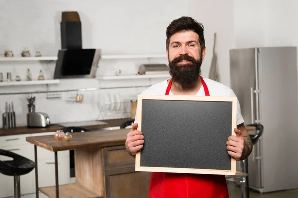 Copy space at his board. Man chef with board, copy space. Mature male. Bearded man cook. Restaurant or cafe menu. Advertisement. Hipster in kitchen. Bearded man in red apron. Ready to cook