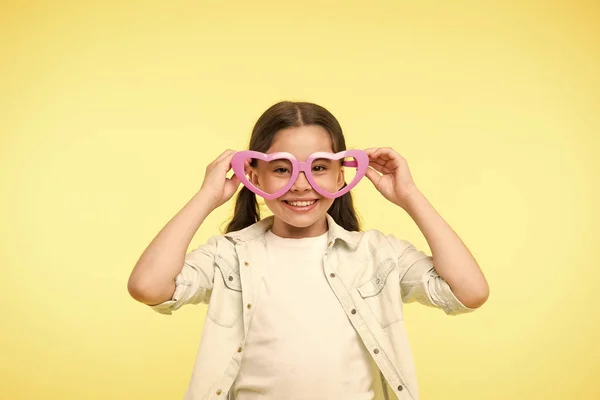 Gelukkig kind draagt een hartvormige bril op een gele achtergrond. Klein meisje glimlach in mode-accessoire. Zomer mode look. Funky stijl schoonheid. Jong en zelfverzekerd. Valentijnsdag. Ik hou van je. — Stockfoto