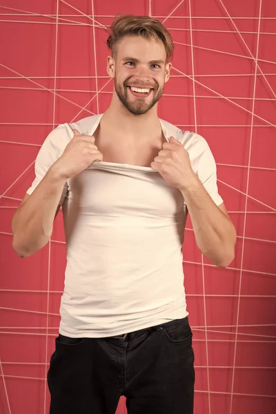 Guy bearded and attractive tearing off shirt. Man with bristle on happy face undressing, pink background. Man with beard or unshaven guy looks handsome and well groomed. Masculinity concept