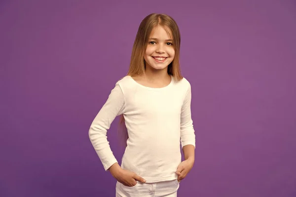 Маленькая девочка улыбается на фиолетовом фоне. Ребенок улыбается с длинными светлыми волосами на фиолетовом фоне. Детская модель в повседневной рубашке. Модный стиль и тренд. Счастливого детства и ухода за детьми — стоковое фото
