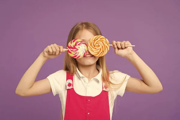 Lustiges Kind mit Lutschern auf violettem Hintergrund. kleines Kind lächelt mit Bonbons auf Stöcken auf lila Hintergrund. Mädchen lächelt mit wirbelnden Karamellen. Essen und Dessert. Schönheit mit Süßigkeiten. Ernährung und Diät — Stockfoto