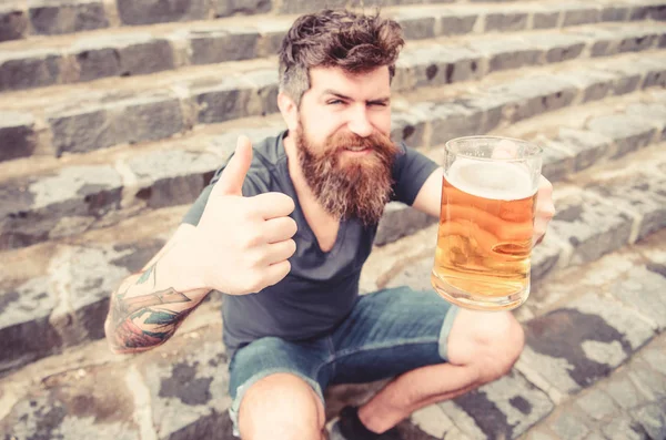 Homem com barba e bigode segura vidro com cerveja e mostra polegar para cima, fundo escadas de pedra. Um tipo a descansar com cerveja, desfocado. Hipster na cara alegre bebe cerveja ao ar livre. Sexta-feira relaxar conceito — Fotografia de Stock