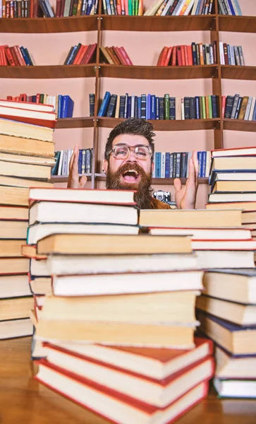 Scientific discovery concept. Man on excited face between piles of books in library, bookshelves on background. Teacher or student with beard wears eyeglasses, sits at table with books, defocused