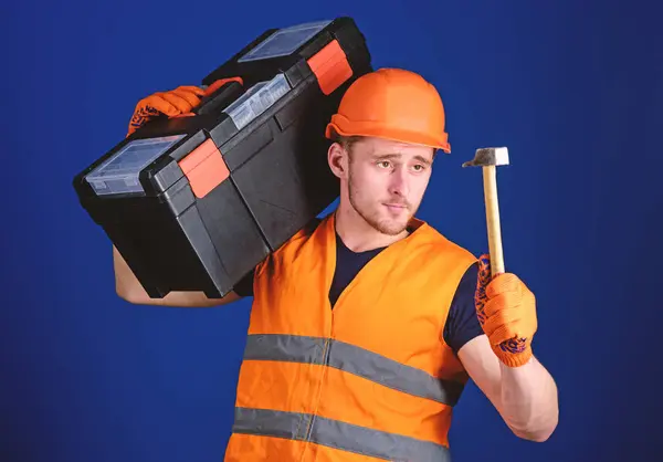 Homem de capacete, chapéu duro carrega caixa de ferramentas e segura martelo, fundo azul. Conceito de faz-tudo. Trabalhador, reparador, reparador, construtor na cara calma carrega caixa de ferramentas no ombro, pronto para o reparo — Fotografia de Stock