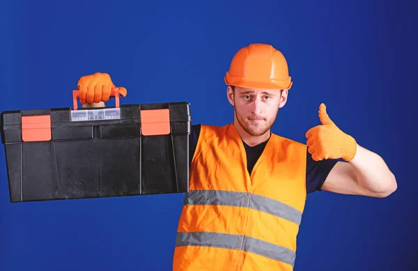 Worker, repairer, repairman, builder on confident face carries toolbox on shoulder. Repair consultation concept. Man in helmet, hard hat holds toolbox and shows thumb up gesture, blue background — Stock Photo, Image