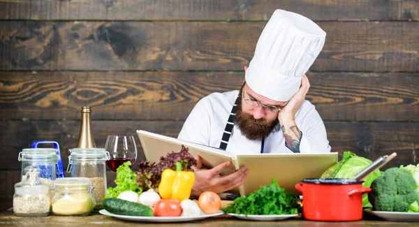 Man bearded hipster read book recipe near table fresh vegetables. Culinary arts. Recipe to cook healthy food. Experienced chef cooking excellent dish. This recipe is just perfect. Vegetarian recipe