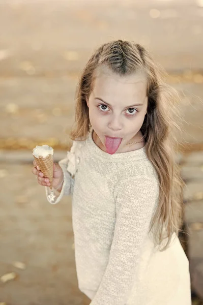 Chica goloso en alegre mueca con lengua come helado, fondo claro. Niña con cono de helado en la mano. El verano trata el concepto. Niño goloso con helado blanco en cono de gofre — Foto de Stock