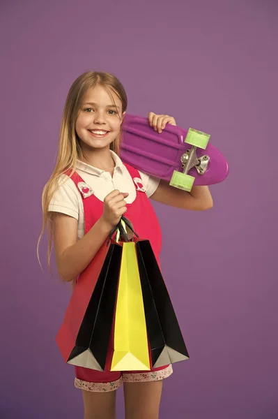 Petit sourire d'enfant avec des sacs en papier et planche à roulettes sur fond violet. Fille heureuse avec planche à roulettes et sacs à provisions. Shopping et vendredi noir. Sport et jeux actifs. Joyeux shopaholic fille . — Photo