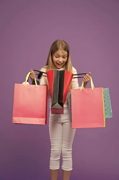 Kind kijk in boodschappentassen op violette achtergrond. Weinig shopaholic glimlach verrast met papieren zakken. Jeugd. Winkelen en verkoop. Shopper meisje met paperbags. Wat een verrassing. Grote verkoop en winkelen — Stockfoto