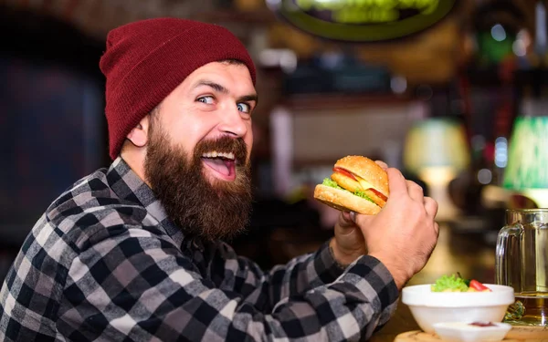 Um homem faminto de Hipster come hambúrguer. Um homem com barba come hambúrguer. Homem barbudo hipster brutal sentado no balcão do bar. Comida de alta caloria. Comida de batota. Delicioso conceito de hambúrguer. Desfrute do sabor de hambúrguer fresco — Fotografia de Stock