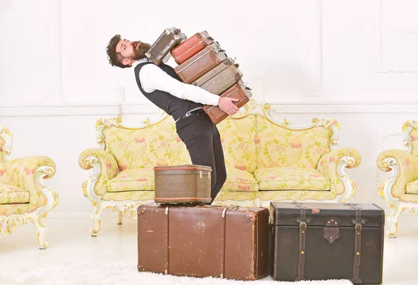 Man with beard and mustache wearing classic suit delivers luggage, luxury white interior background. Macho, elegant porter carries heavy pile of vintage suitcases. Butler and service concept