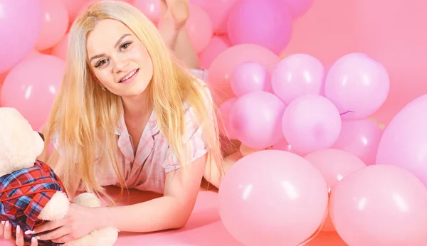 Blonde on smiling face relaxing with teddy bear toy. Girl in pajama, domestic clothes lay near air balloons, pink background. Birthday girl concept. Woman cute celebrate birthday with balloons