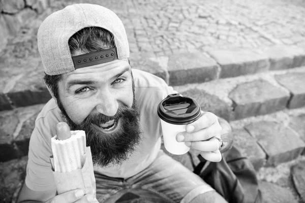 Homme barbu profiter de la nourriture de rue fond urbain. Saucisse savoureuse dans un pain rôti. Concept de culture alimentaire urbaine. Hipster manger hot dog tout en s'asseyant sur les escaliers à l'extérieur. Hipster profiter hot dog et boire tasse en papier — Photo